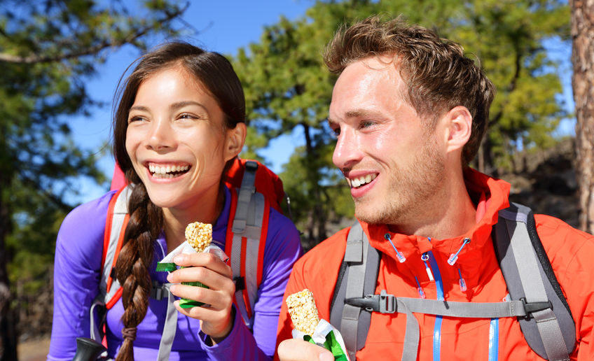 32707644 - couple eating muesli bar hiking. happy people enjoying granola cereal bars living healthy active lifestyle in mountain nature. woman and man hiker sitting laughing during hike.